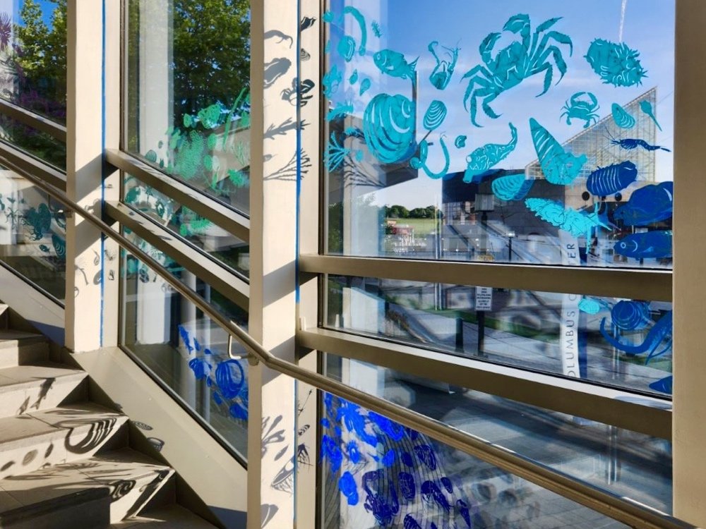 Art installation by April Flanders. Silhouettes of marine invertebrates in vibrant blues and greens appear to float across the sunlit windows of the Colwell Center.